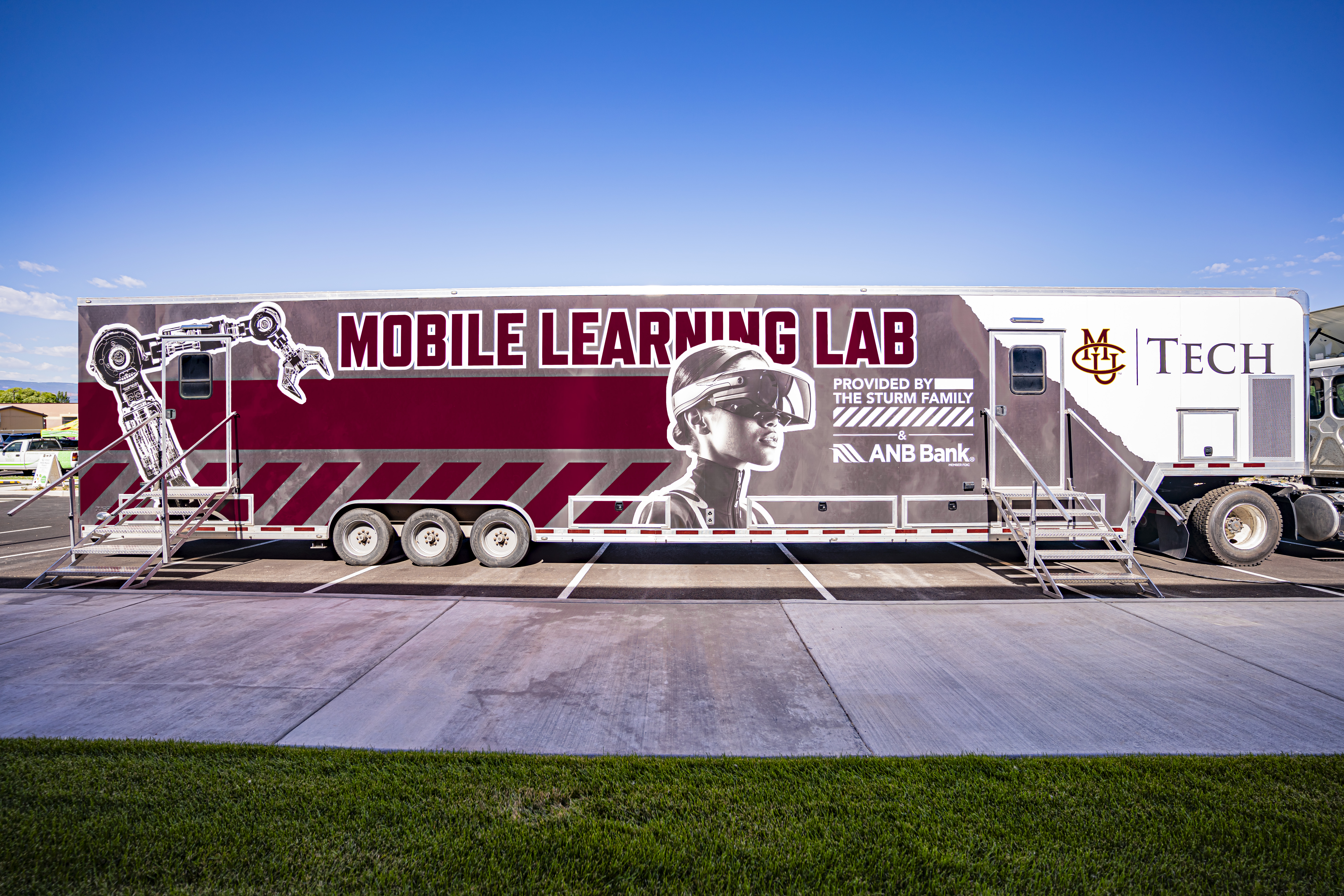 The image shows a large mobile trailer parked in a paved parking lot with a clear blue sky overhead. The trailer is primarily maroon and white and is branded as the “Mobile Learning Lab.”  On the far left side of the trailer, there is a large graphic of a robotic arm in black and white. In the center, there is an image of a person wearing protective safety goggles, looking upward, symbolizing innovation and hands-on learning. Beside this image, bold maroon letters read: “Mobile Learning Lab.” Beneath the text, it states: “Provided by The Sturm Family & ANB Bank.”  On the far right of the trailer, the CMU Tech logo is prominently displayed in maroon and gold. The trailer sits on three axles with metal steps providing access to doors at both ends. The setting includes a green lawn in the foreground and a sidewalk running along the front of the trailer. The overall design emphasizes technology, mobility, and community education.