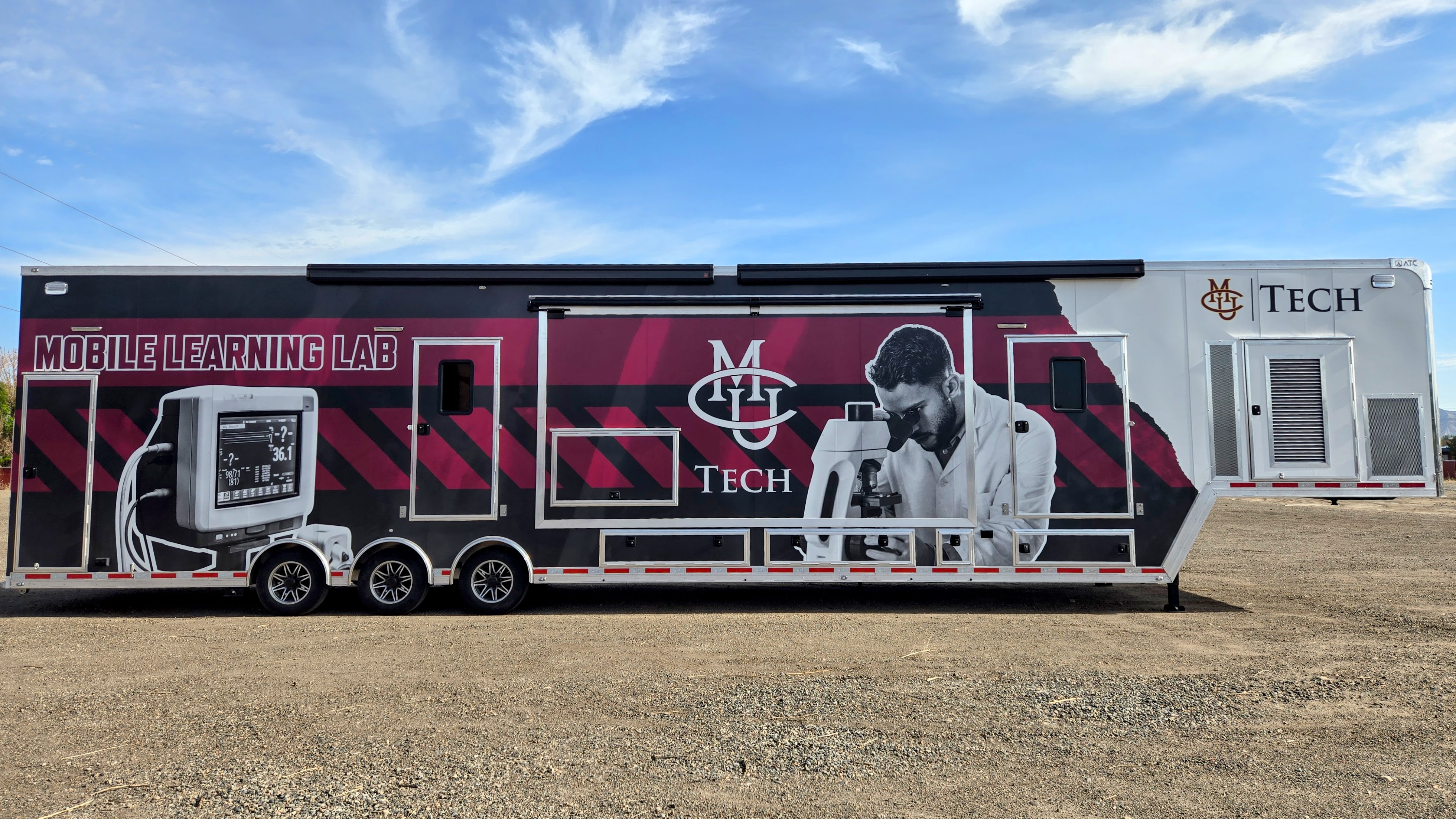 The image shows a large mobile trailer parked in a dirt parking lot with a clear blue sky overhead. The trailer is primarily maroon and white and is branded as the “Mobile Learning Lab.”On the far left side of the trailer, there is a large graphic of a vital signs monitor in black and white. In the center, there is an image of a person wearing a lab coat, looking downward into a microscope, symbolizing innovation and hands-on learning. Beside this image, bold maroon letters read: “Mobile Learning Lab.”On the far right of the trailer, the CMU Tech logo is prominently displayed in maroon and gold. The trailer sits on three axles with metal steps providing access to doors at both ends.The overall design emphasizes technology, mobility, and community education.